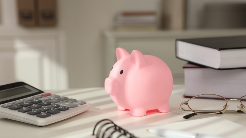 Kasica prasica na stolu pored digitrona, knjiga i naočara kao simbol kontrole finansija u biznisu