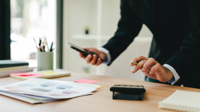 Osoba u odelu proverava troškove na digitronu uz papire i telefon kao deo finansijskog planiranja u biznisu