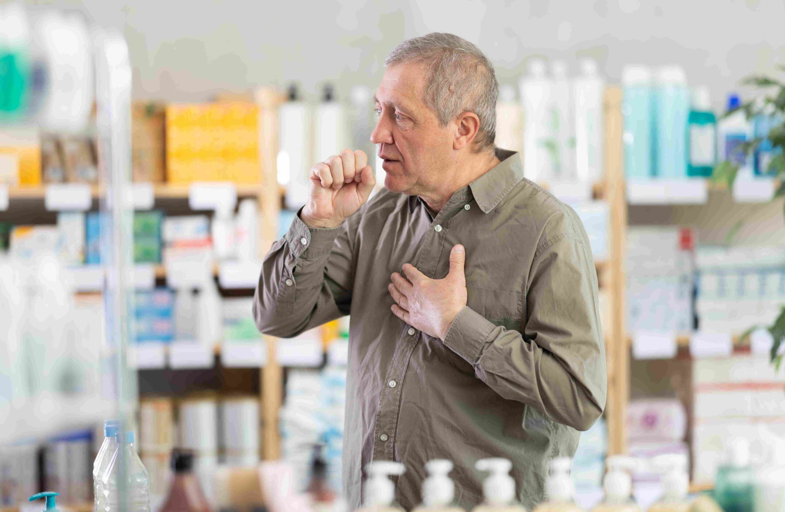 Stariji muškarac kašlje i drži ruku na grudima u apoteci