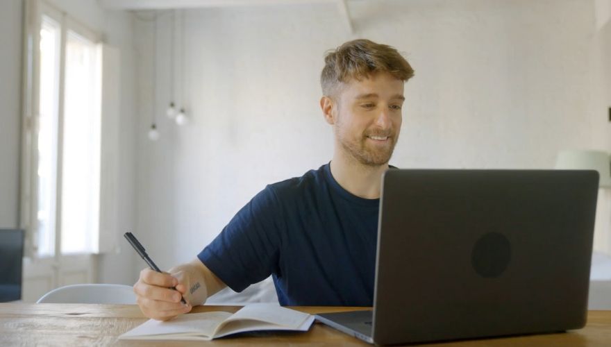 Mladi muškarac prati online nastavu na laptopu i zapisuje beleške u svesku, usvajajući nova znanja i veštine kroz program prekvalifikacije.